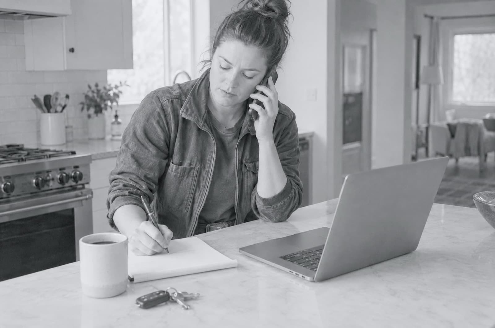 A business owner on the intake call at her kitchen counter, taking notes beside a laptop