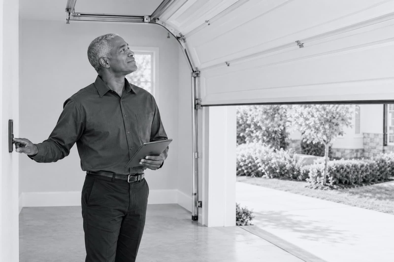 A man with a tablet pressing the wall control of an open garage door, looking up at it.