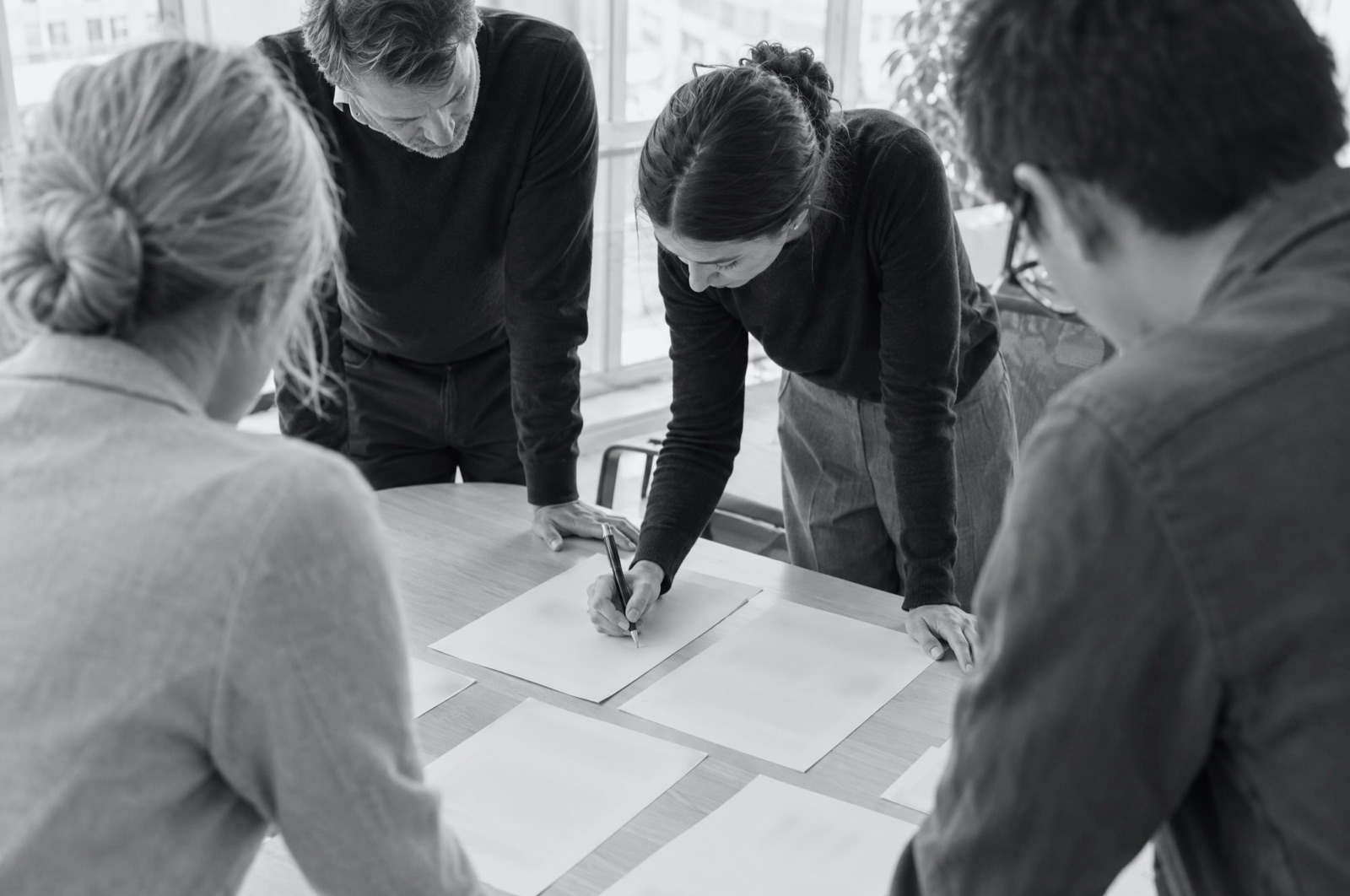 Four managers standing around a table of papers, one leaning in to sign a page