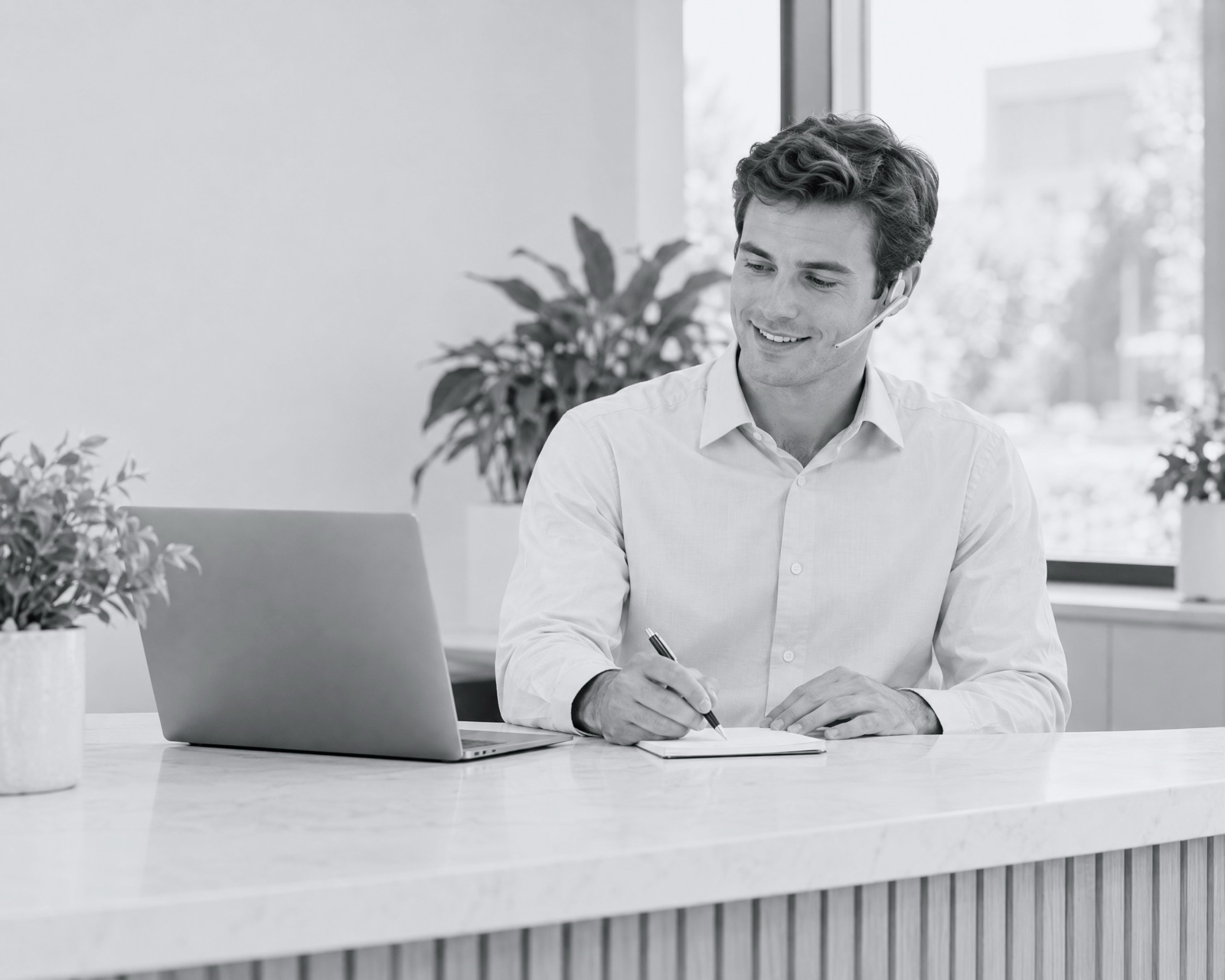 An office coordinator on a headset call at a bright front desk, taking notes.
