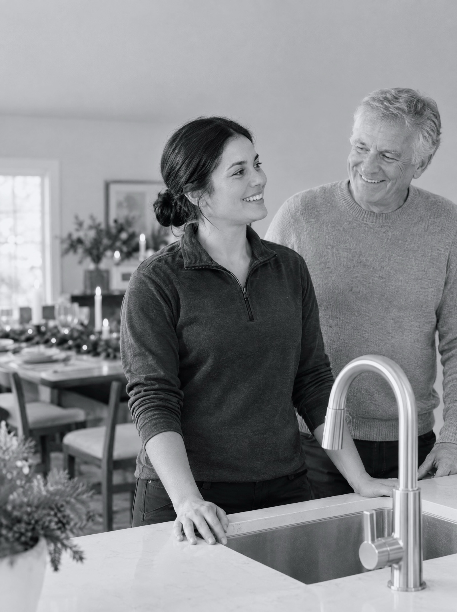 A technician at the sink of a tidy holiday kitchen, the host smiling beside her, a set table behind
